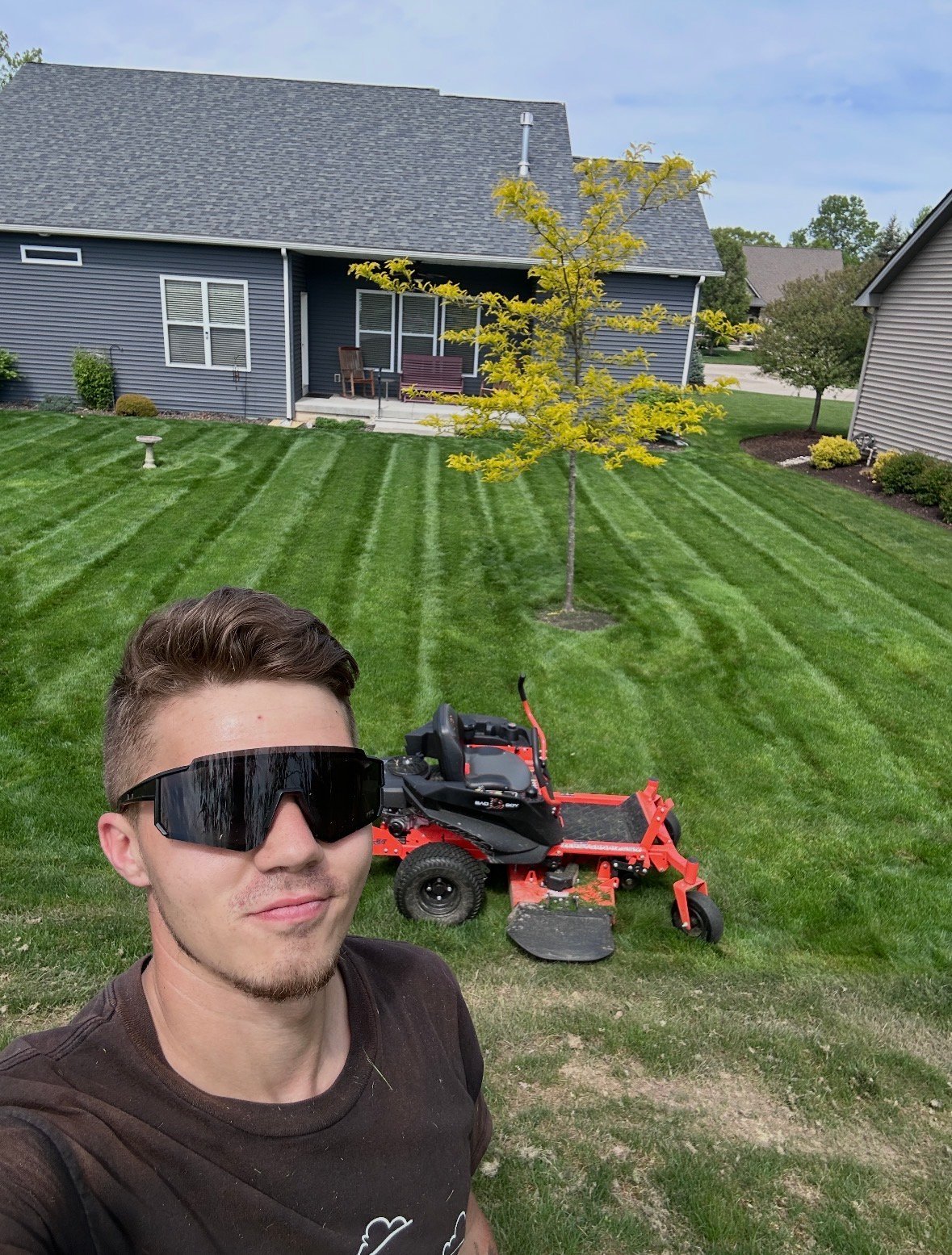 Man wearing dark sunglasses posing in front of red zero-turn mower on striped lawn with house in background