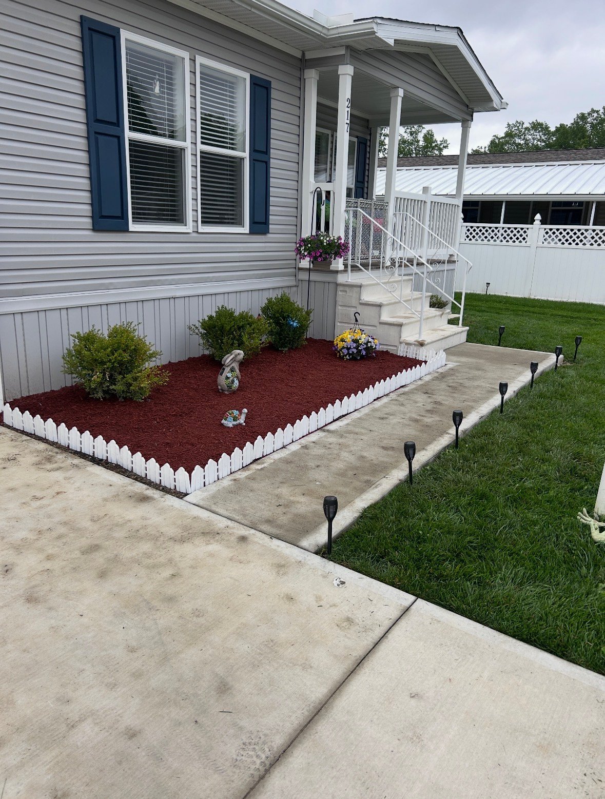Front exterior of a mobile home with gray siding, blue shutters, white porch, red mulch garden bed with landscaping, and manicured lawn