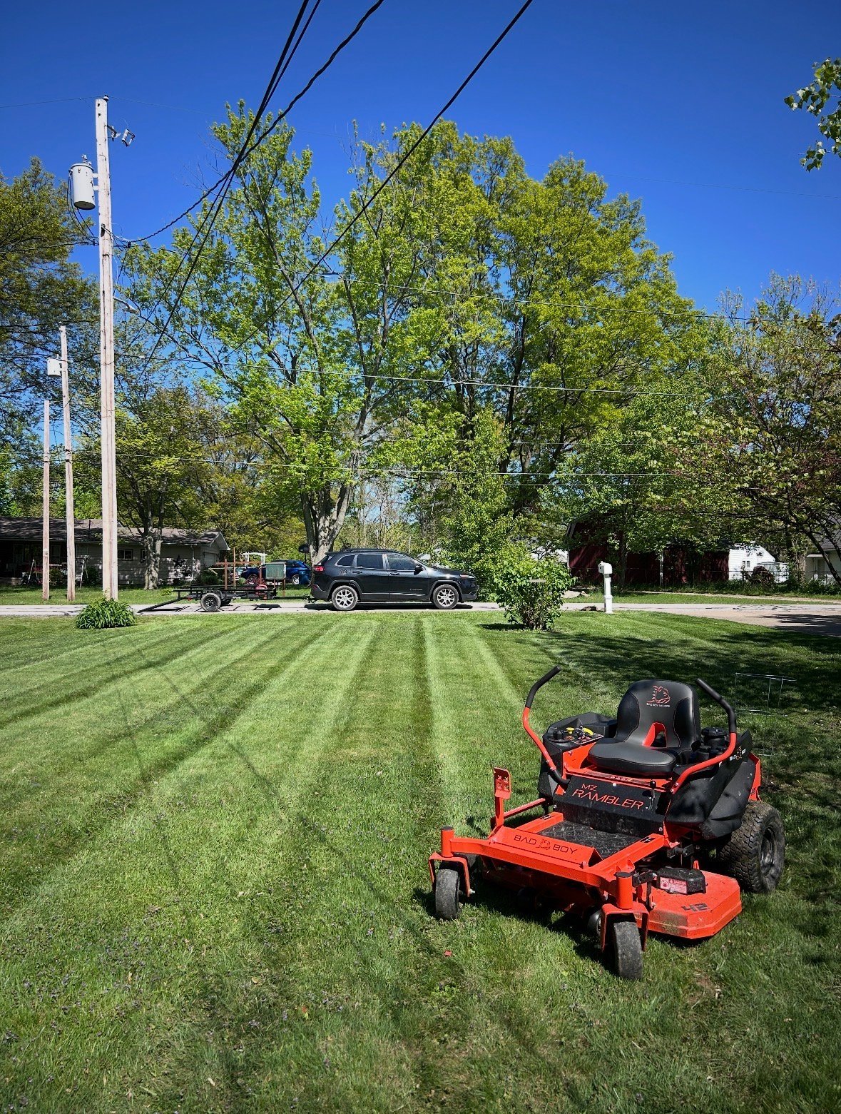 Red zero-turn mower on freshly striped lawn with utility pole, trees, and parked vehicles in background under clear blue sky